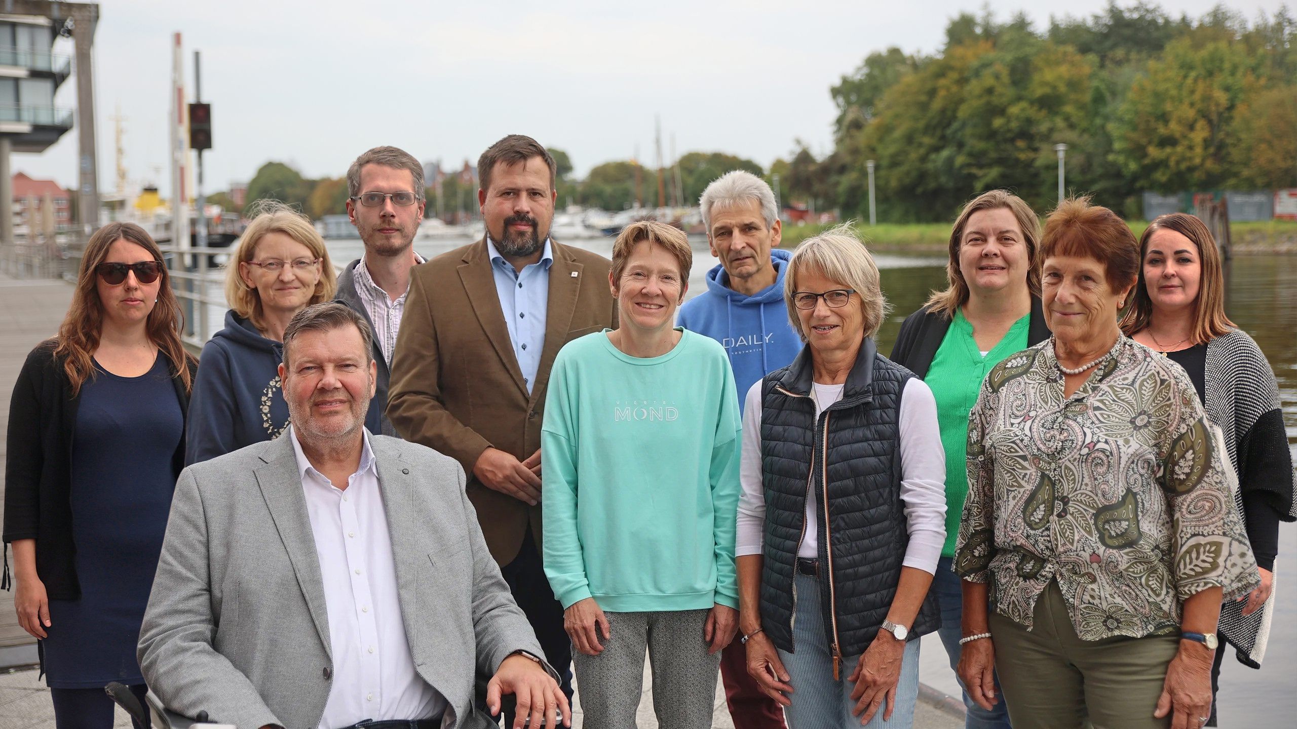 Das Gruppenbild zeigt den Trägerkreis. Sieben Personen (von links nach rechts, Sabine Hilbers, Stefan Kamer, Sonja Renken-Muehlbacher, Matthias Meyer, Anne Groeneweg, Sandra Meyer und Elfriede Wilts) stehen am Emder Delft. Ein Mann (Jörn Malanowski) sitzt im Rollstuhl, trägt einen grauen Anzug und ein weißes Hemd. Die Frauen und Männer stehen hinter ihm, eine trägt ein grünes Oberteil, eine eine schwarze Weste und die anderen zwei hellgrüne und gemusterte Kleidung.
